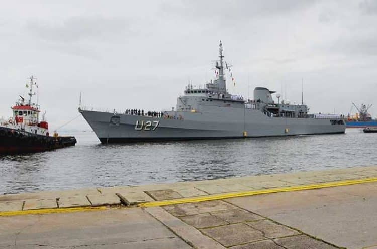 A navy ship docked in the water next to a tug boat.
