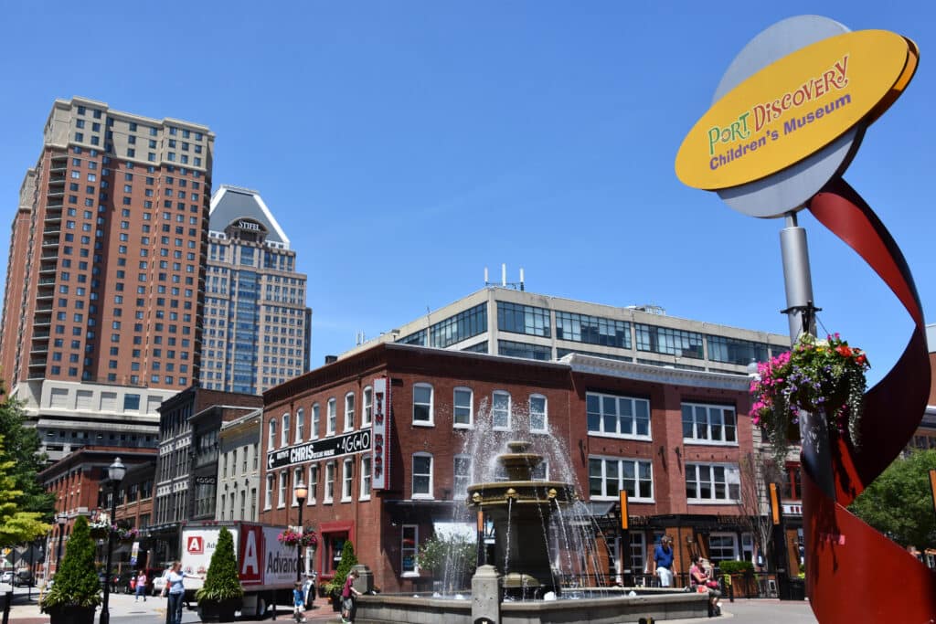 Outdoor scene with Port Discovery Children's Museum sign, red brick buildings, a fountain, and tall office buildings under a clear blue sky.
