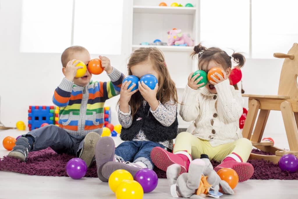 Three children sitting on a rug hold colorful plastic balls to their faces, surrounded by toys in a playroom.