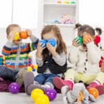 Three children sitting on a rug hold colorful plastic balls to their faces, surrounded by toys in a playroom.
