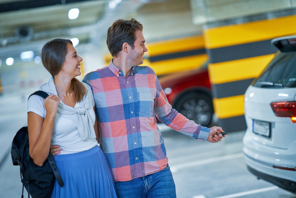 A man and woman walking together in a parking garage, with the man holding a car key and pointing toward a white SUV.