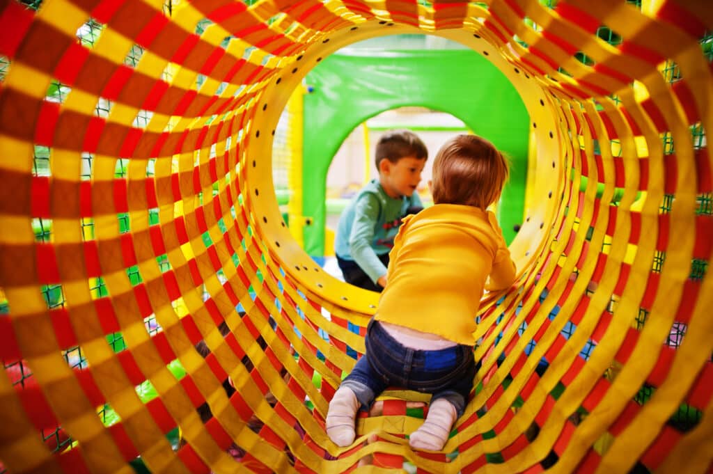Children play inside a colorful, woven tunnel in a play area.