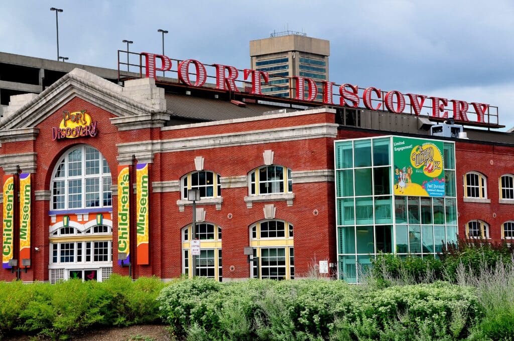 Exterior of Port Discovery Children's Museum, a red-brick building with glass facade and colorful banners, under a cloudy sky.
