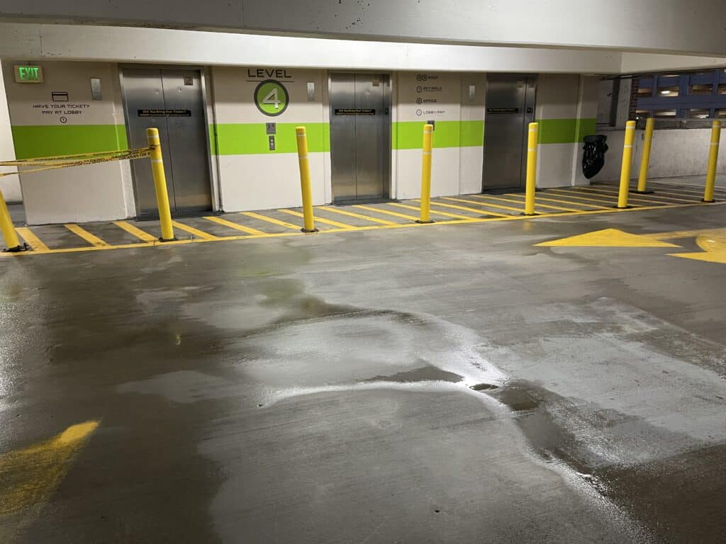 Three elevators on level 4 of a parking garage, with yellow safety poles and wet floor; directional arrow painted on the concrete.