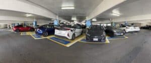 Wide view of a multi-level parking garage with several parked cars, including sedans and electric vehicles, in marked parking spaces under fluorescent lighting.