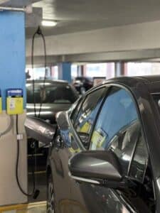A car is parked in a parking garage, plugged into an electric vehicle charging station mounted on a blue pillar.