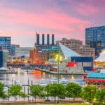 Baltimore Inner Harbor at sunset, featuring modern buildings, the National Aquarium, waterfront, boats, and colorful sky.