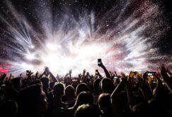A crowd of people at a concert holds up phones and hands as bright stage lights and confetti fill the air.