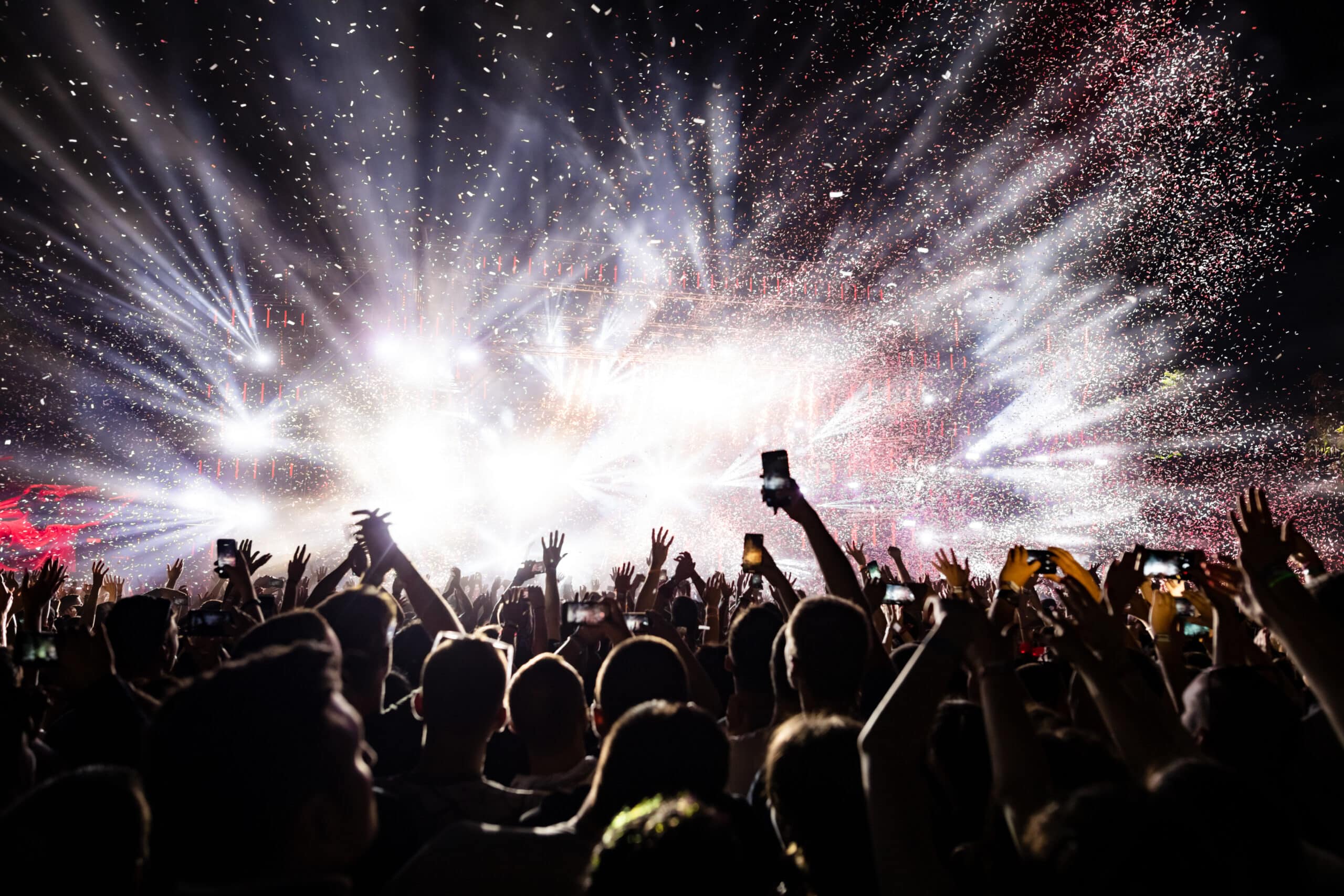 A crowd of people at a concert holds up phones and hands as bright stage lights and confetti fill the air.
