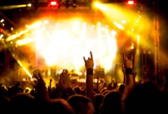 A crowd at a concert raises their hands, some making the rock and roll gesture, with bright yellow stage lights illuminating the scene.