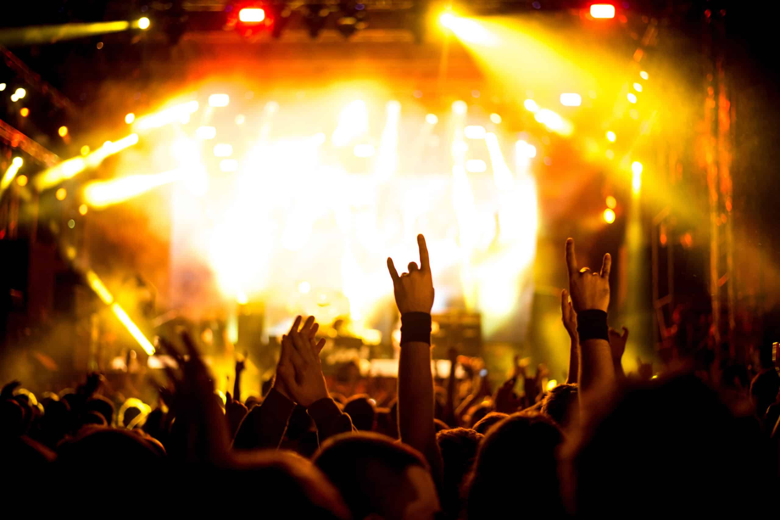 A crowd at a concert raises their hands, some making the rock and roll gesture, with bright yellow stage lights illuminating the scene.