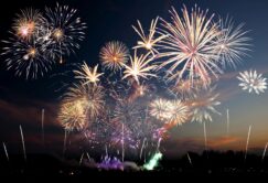 A large display of colorful fireworks explodes in the night sky above a dark landscape, with bright bursts and trails lighting up the scene.