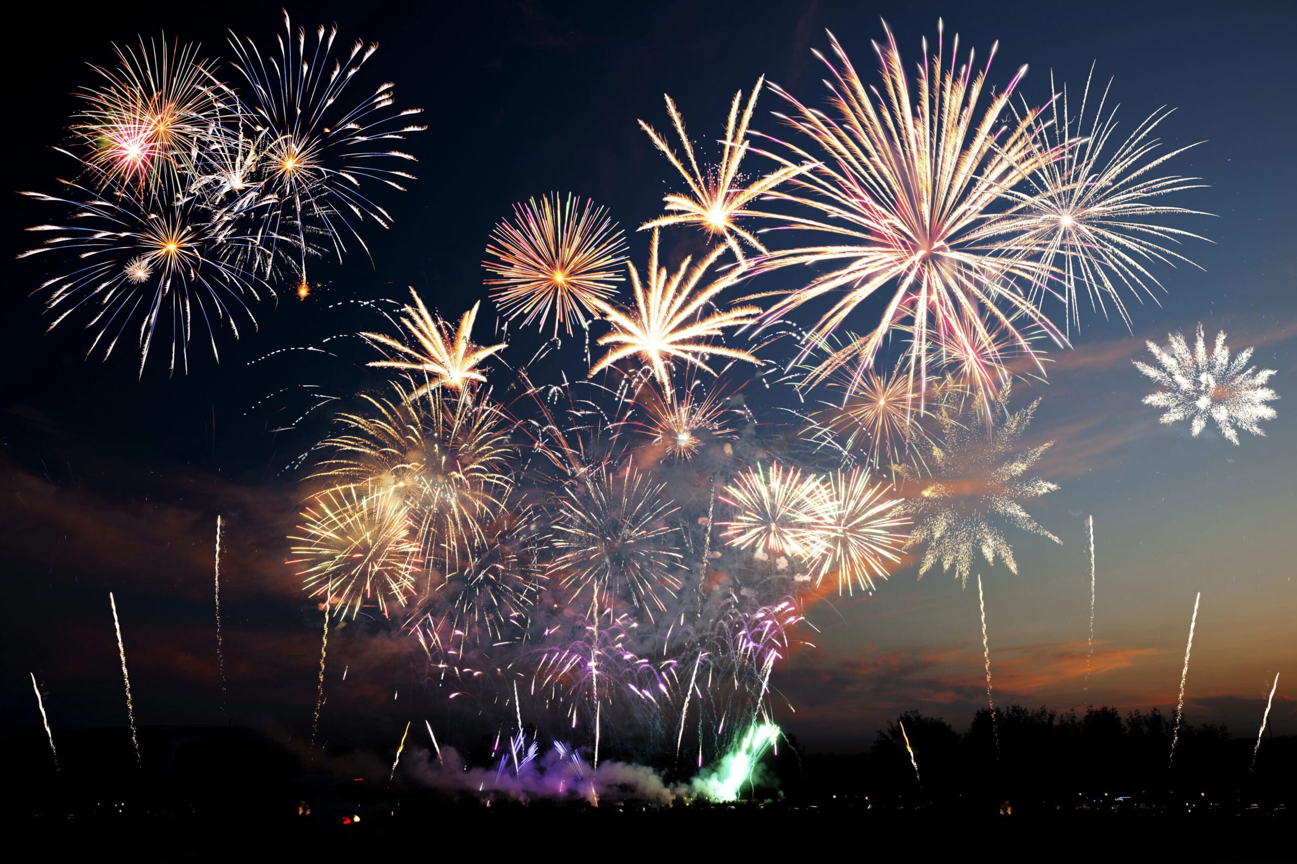 A large display of colorful fireworks explodes in the night sky above a dark landscape, with bright bursts and trails lighting up the scene.