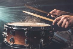 Close-up of a person striking a snare drum with drumsticks, causing particles to rise from the drumhead in dramatic lighting.