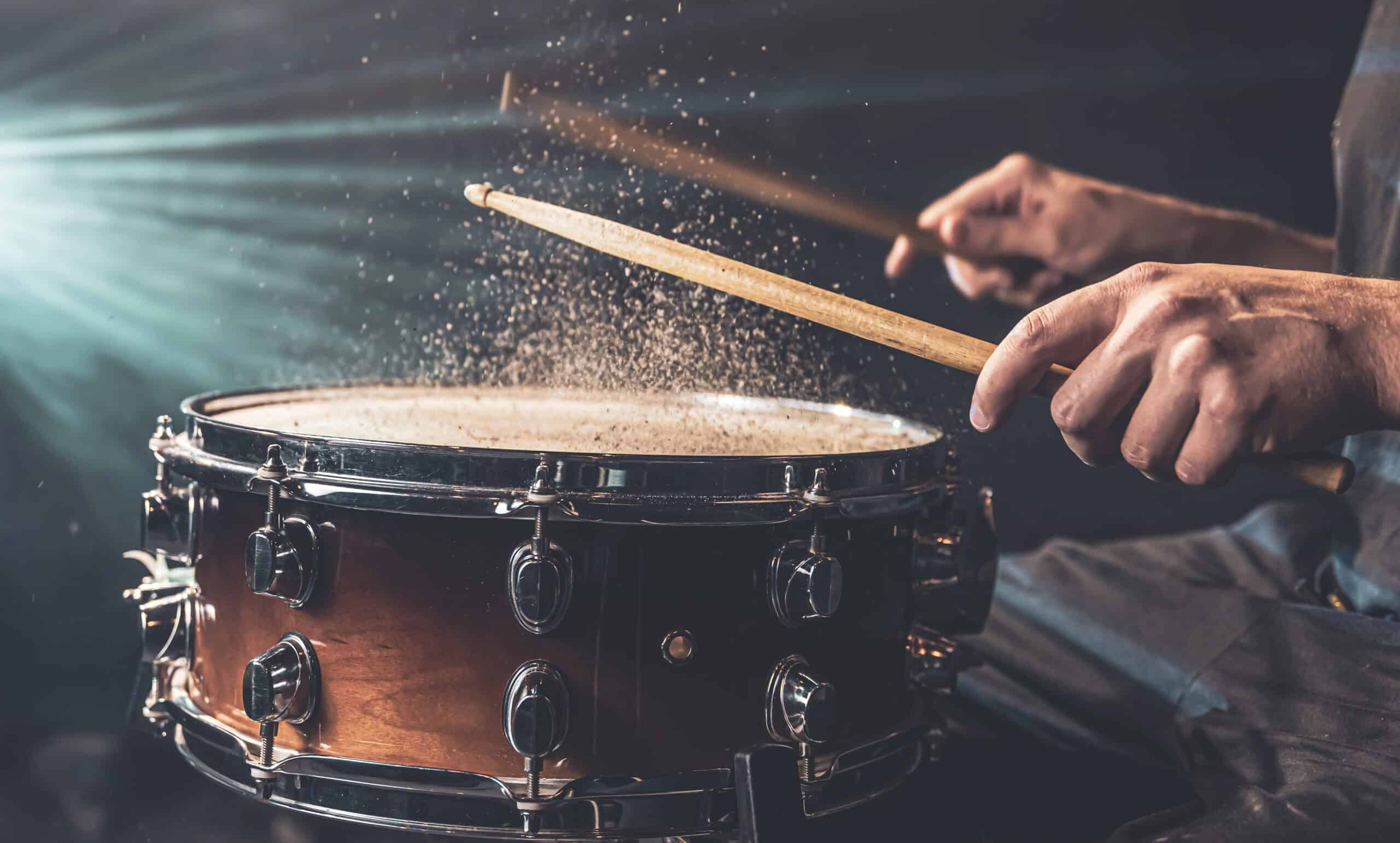 Close-up of a person striking a snare drum with drumsticks, causing particles to rise from the drumhead in dramatic lighting.