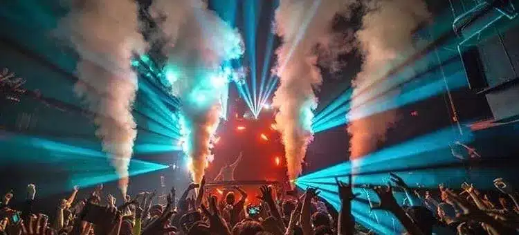 Crowd at a concert with raised hands, stage lights, smoke effects, and a performer visible in the center under bright beams.