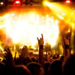 Crowd at a concert raising hands and making rock-and-roll gestures, illuminated by bright yellow stage lights.