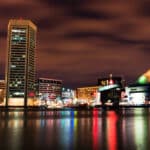 Night view of a city waterfront with tall buildings and a glass pyramid structure, colorful lights reflected on calm water under a cloudy sky.