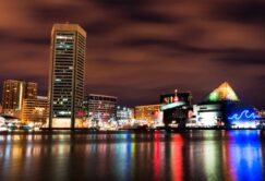 Night view of a city waterfront with tall buildings and a glass pyramid structure, colorful lights reflected on calm water under a cloudy sky.