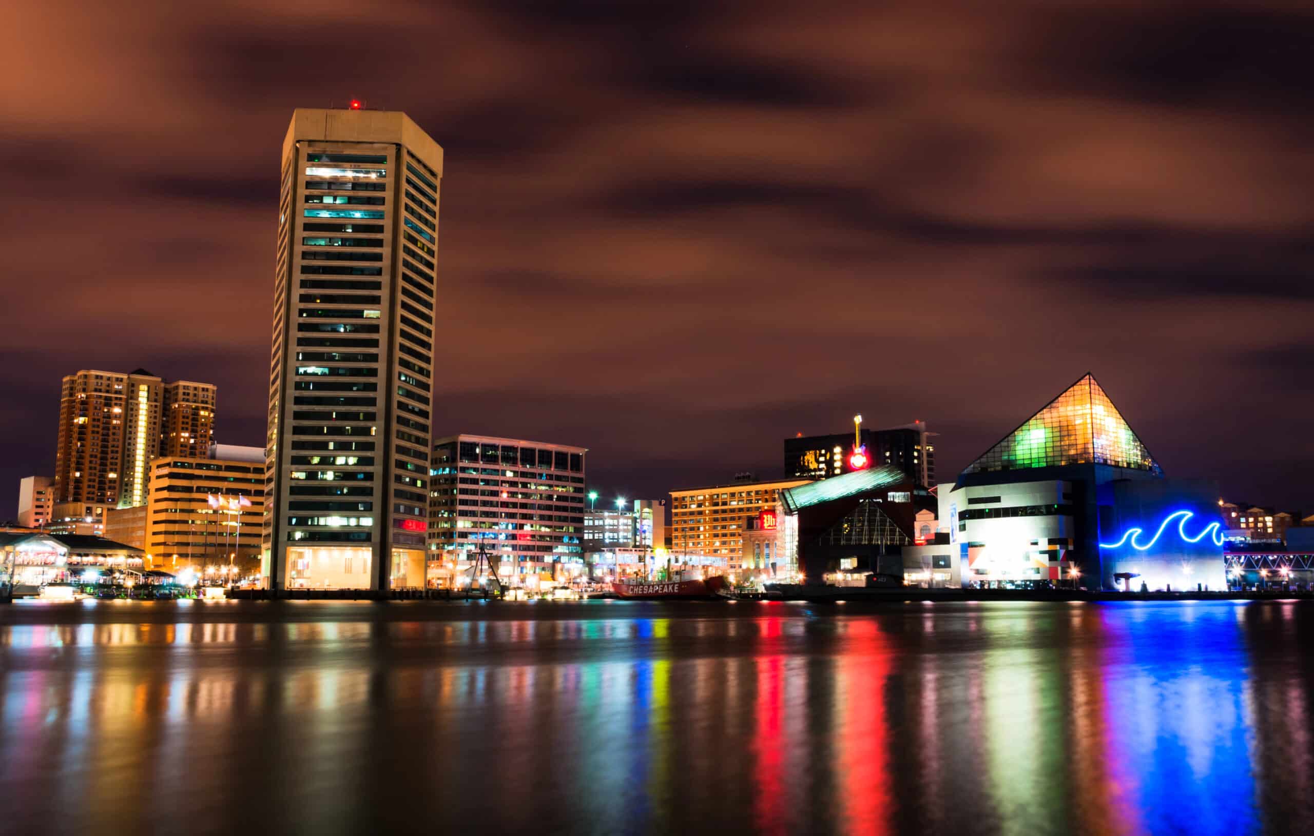 Night view of a city waterfront with tall buildings and a glass pyramid structure, colorful lights reflected on calm water under a cloudy sky.
