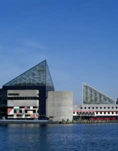 A modern waterfront building with glass pyramid roofs and geometric concrete walls, seen from across the water under a clear blue sky.
