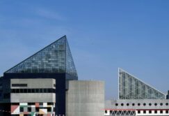 A modern waterfront building with glass pyramid roofs and geometric concrete walls, seen from across the water under a clear blue sky.