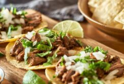 Three tacos topped with shredded meat, chopped onions, and cilantro on a wooden tray, with lime wedges and a bowl of tortilla chips in the background.
