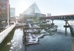 A waterfront scene with snow-covered walkways, a glass pyramid building, bridges, and modern city buildings under bright sunlight.
