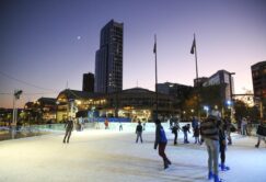 People ice skating at an outdoor rink in a city at dusk, with tall buildings and flags visible in the background under a twilight sky.