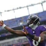 A football player in a purple Baltimore Ravens uniform throws a football on a stadium field with empty blue seats in the background.