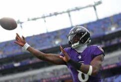 A football player in a purple Baltimore Ravens uniform throws a football on a stadium field with empty blue seats in the background.