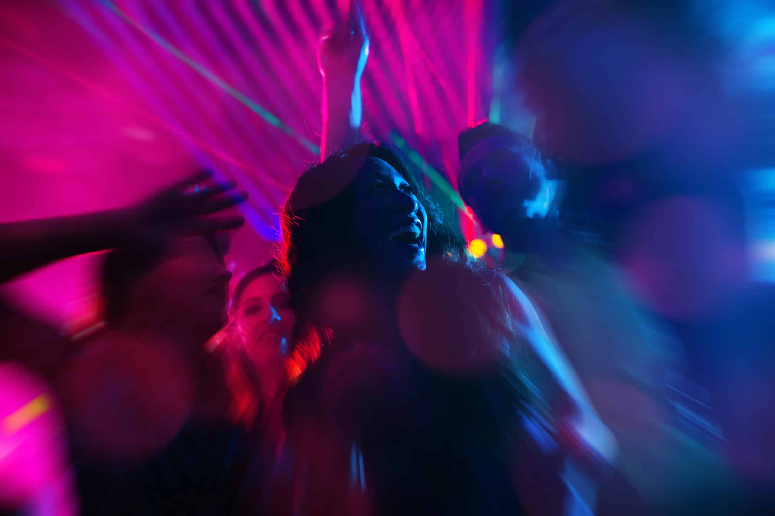 A group of people dance under colorful neon lights at a party, with blurred motion and vibrant pink and blue hues creating a dynamic atmosphere.