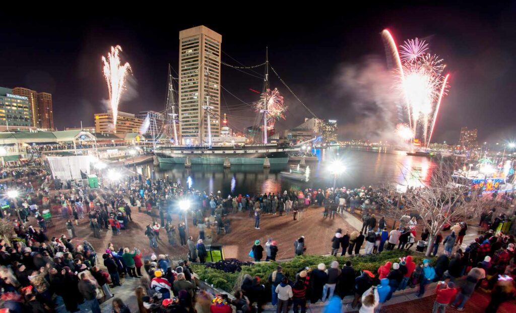 A large crowd gathers at a waterfront area at night to watch fireworks lighting up the sky, with tall buildings and a historic ship in the background.