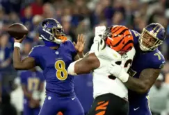 A football quarterback in a blue uniform prepares to throw the ball as a teammate blocks a defender in a white and orange uniform during a game.