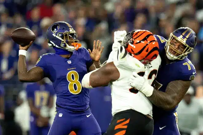 A football quarterback in a blue uniform prepares to throw the ball as a teammate blocks a defender in a white and orange uniform during a game.