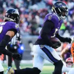 Two Baltimore Ravens football players, Gilman and Van Noy, in purple uniforms run with the ball during a game against the Cincinnati Bengals, whose players wear orange and white uniforms.