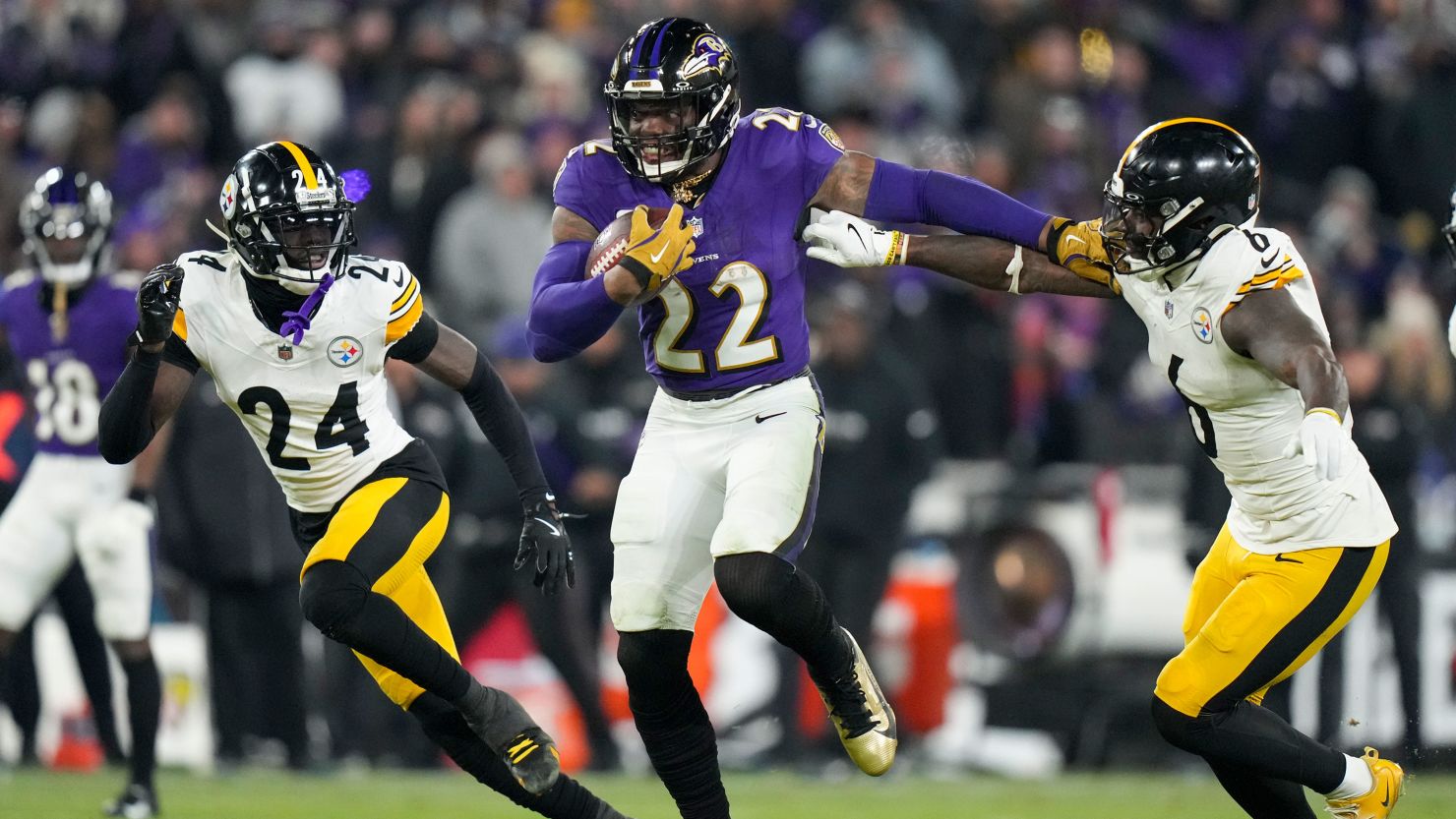 A football player in a purple Ravens uniform runs with the ball while fending off two defenders in white Steelers uniforms during a game.