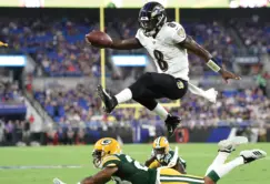 A Baltimore Ravens player leaps over a Green Bay Packers defender during a football game, holding the ball mid-air as other players watch.