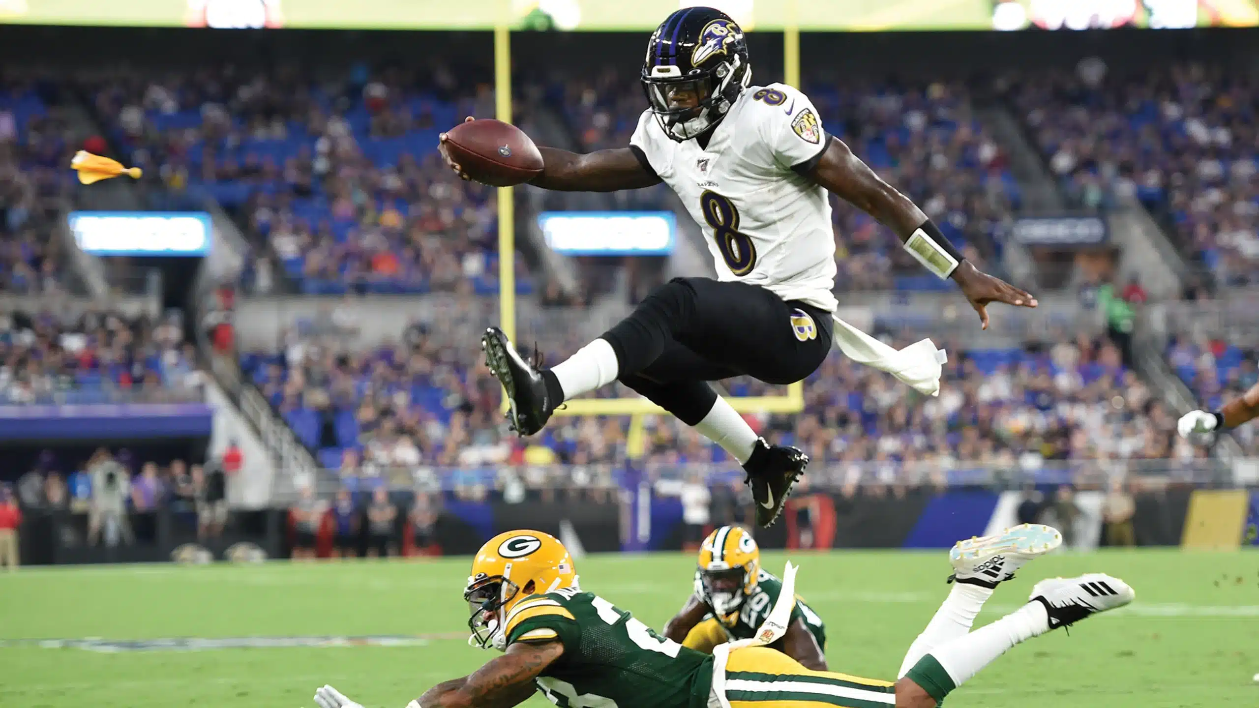 A Baltimore Ravens player leaps over a Green Bay Packers defender during a football game, holding the ball mid-air as other players watch.