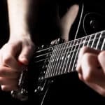Close-up of a person playing an electric guitar, with one hand pressing the strings on the fretboard and the other hand strumming.